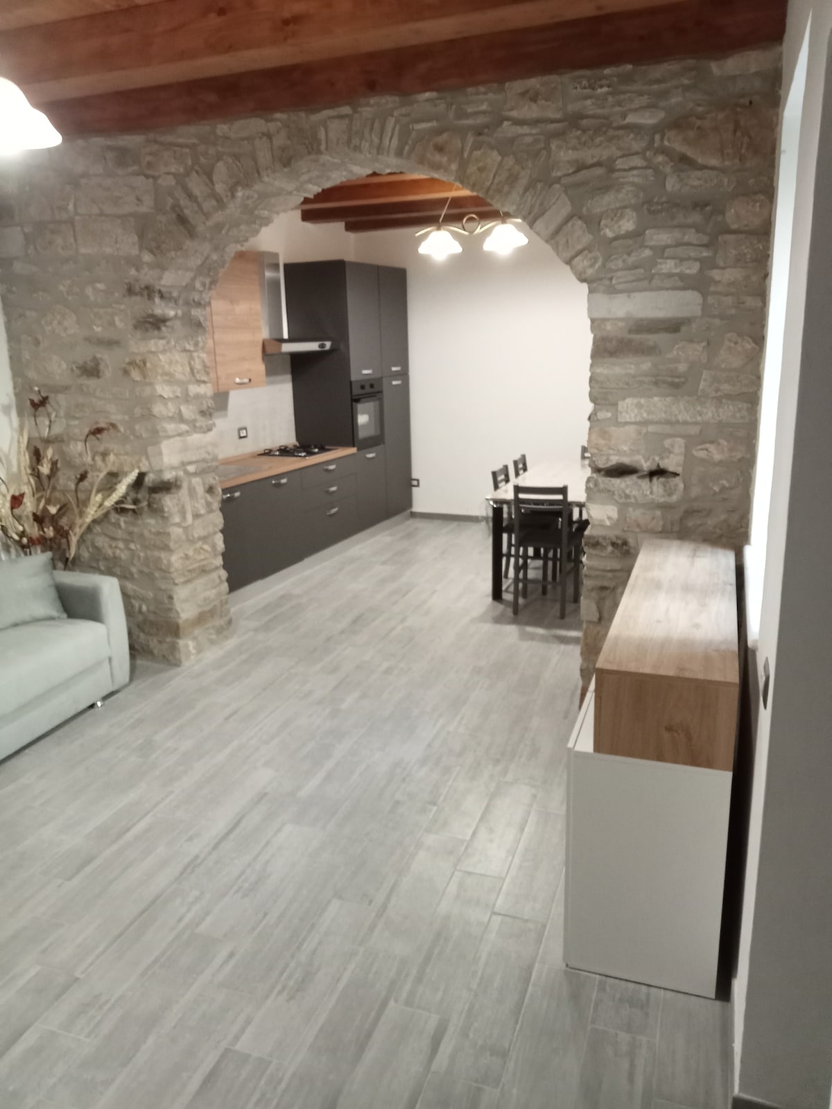 An arched stone entryway leads into a modern kitchen and dining area. The kitchen features sleek cabinetry and appliances, including a stove and oven. A dining table with chairs is positioned beside a neutral-toned sideboard, while wooden beams add character to the ceiling.
