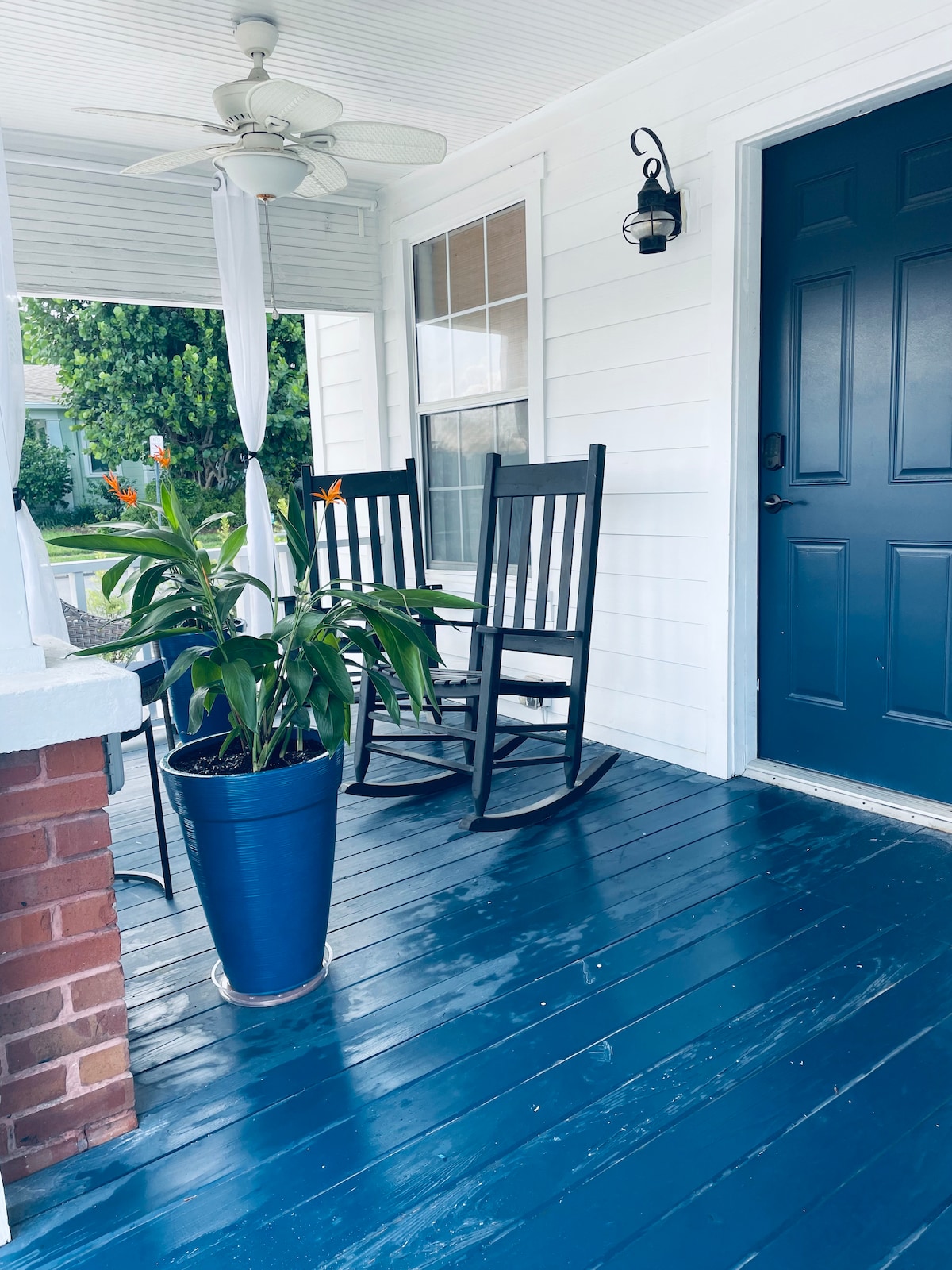 A welcoming porch area features two black rocking chairs positioned beside a large potted plant. Light colors of the wall and elegant blue door contrast with the dark blue floor, creating a calm entrance. A ceiling fan and sheer curtains enhance the outdoor space.