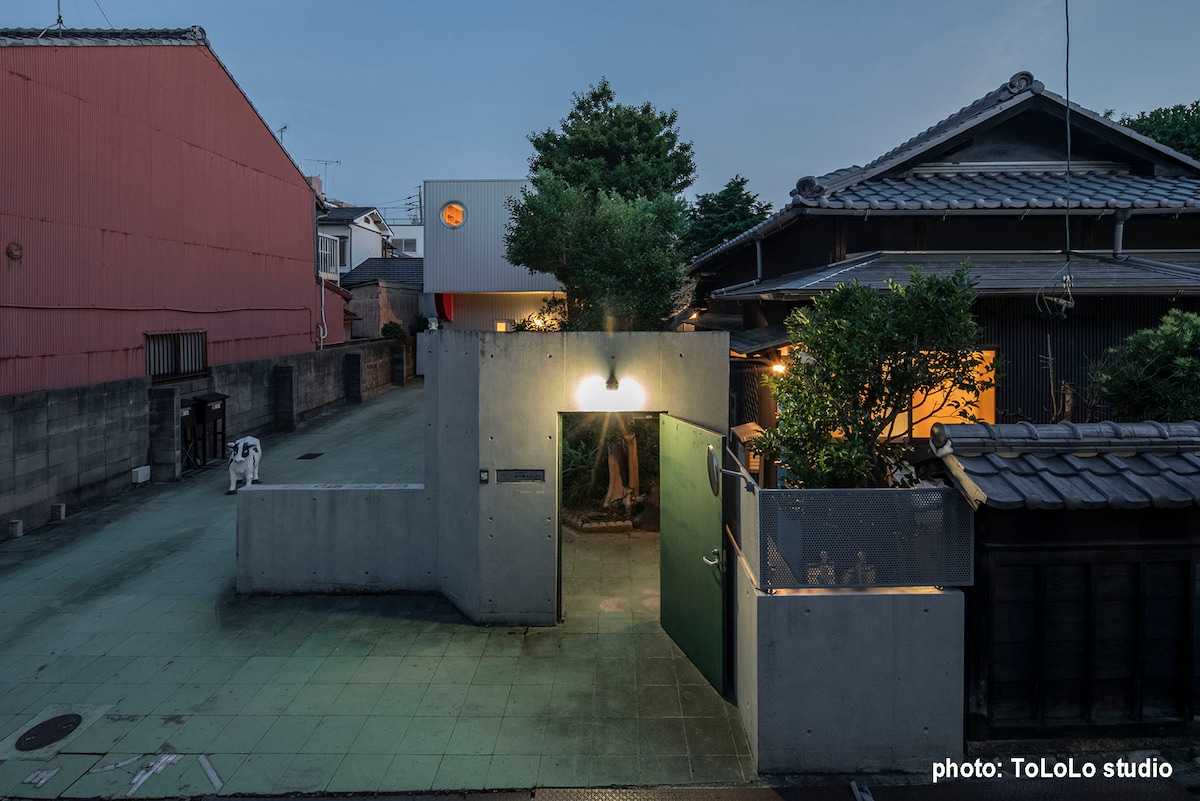 The image shows an entryway to a residential area, featuring a concrete wall with an illuminated entrance. Greenery is visible around the entrance, with traditional Japanese-style buildings in the background. The scene is set during twilight, highlighting the calm ambiance.