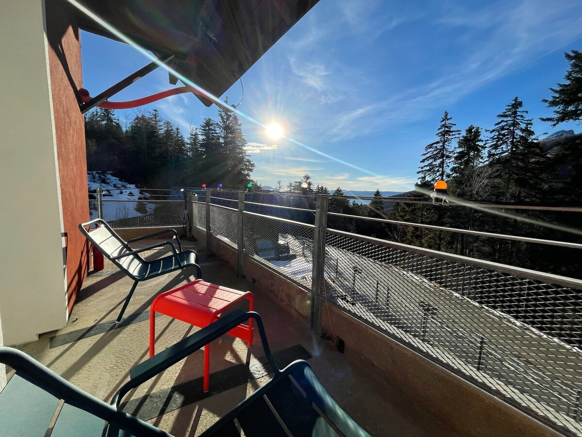 A balcony offers a view of a sunlit mountain landscape, framed by tall trees. Two black chairs and a red table provide comfortable seating. The railing features a chain-link design, allowing for an unobstructed view of the scenery.