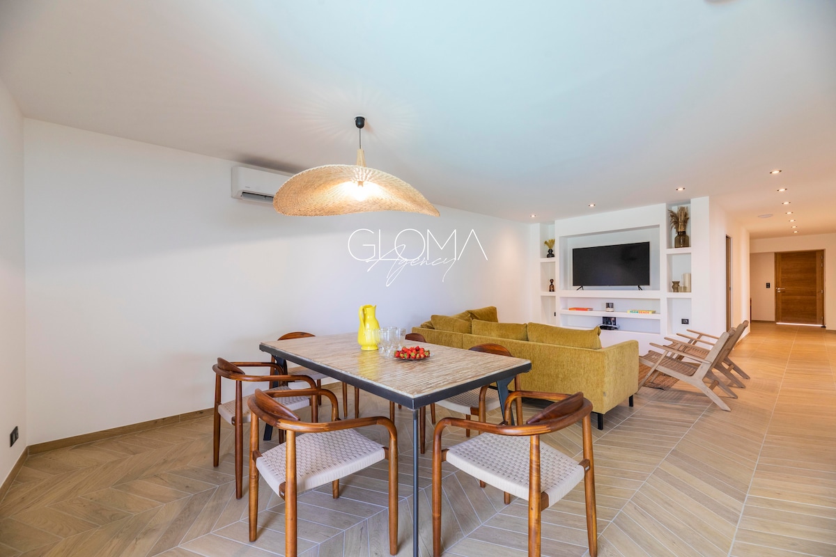 An open-concept living area features a dining table surrounded by six chairs, complemented by a cozy seating arrangement with a large sofa. A modern light fixture hangs above the table, and a wall-mounted television is visible in the background.