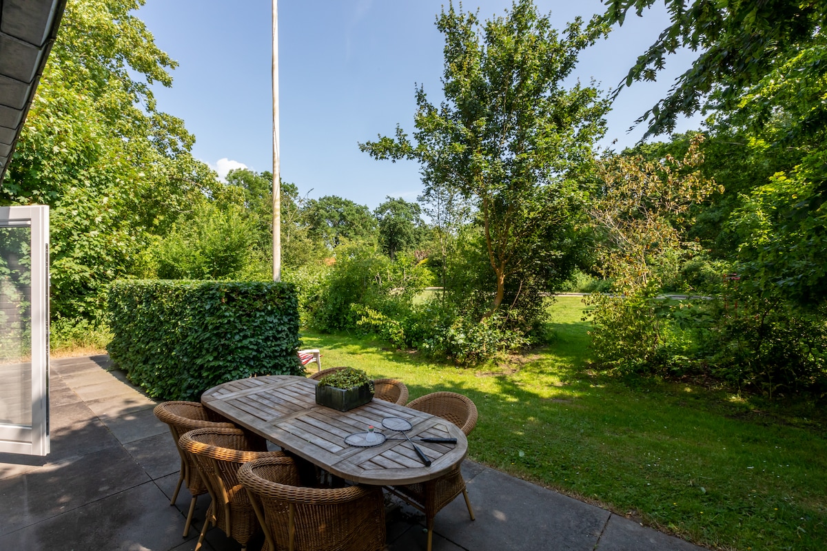 A spacious outdoor dining area features a wooden table surrounded by six wicker chairs. Lush greenery and trees provide a natural backdrop, while a well-maintained lawn is visible in the distance. The area is designed for enjoying meals in a tranquil setting.