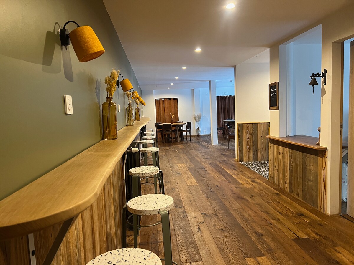 An inviting hallway features a long wooden bar with eclectic stools, creating a warm communal space. Soft lighting illuminates the area, enhancing the natural wood flooring and green accent wall. The background includes rustic dining tables and a cozy seating area.