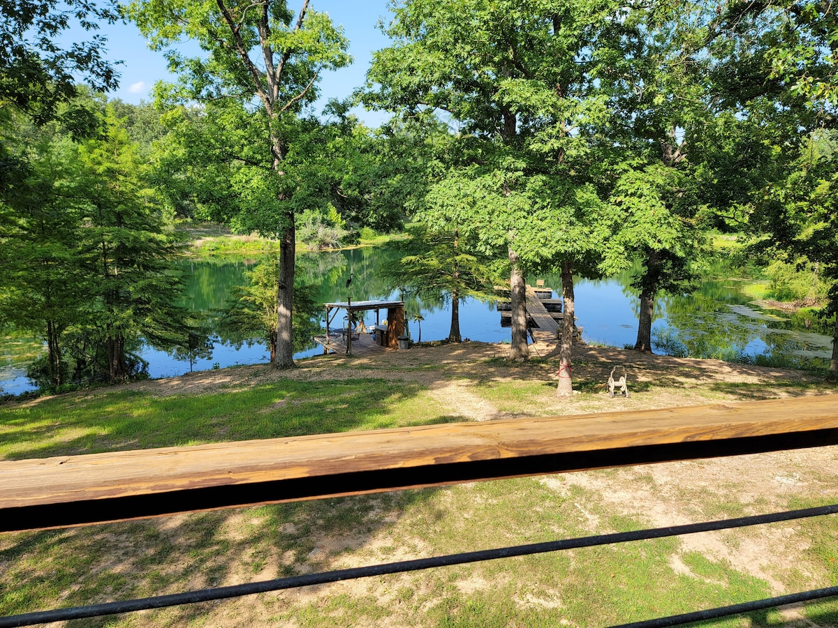 A calm pond is surrounded by lush greenery and trees. Two docks are visible along the water's edge, providing options for fishing or swimming. A small picnic table is positioned under a tree, enhancing the serene outdoor setting.