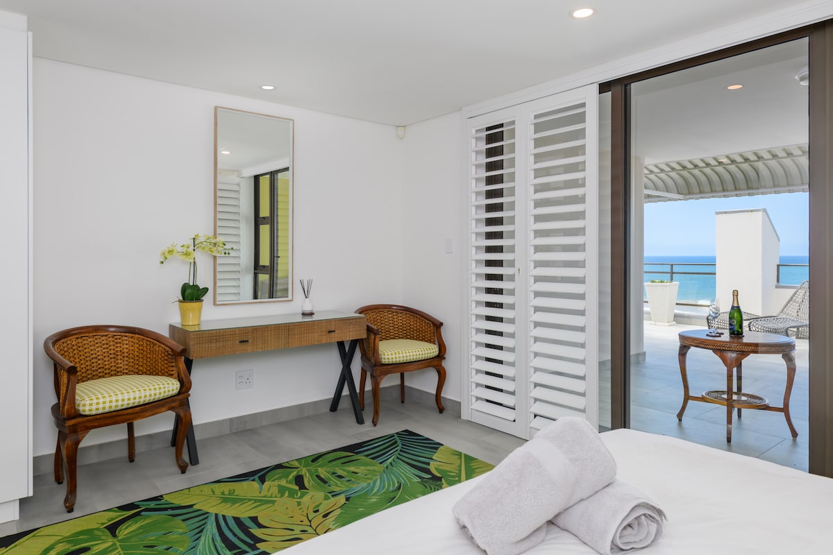 A spacious bedroom features a large bed with fresh towels neatly arranged. A dressing table is positioned against the wall with a large mirror above it. Two comfortable chairs are placed beside the table, and sliding doors open onto a terrace with ocean views.