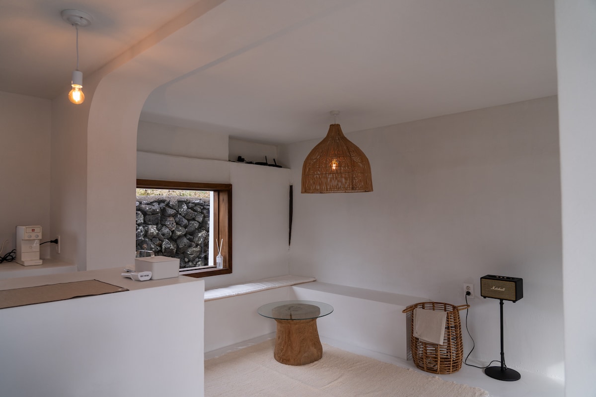 A minimalistic interior space features a round glass coffee table set atop a light-colored rug. A woven pendant lamp hangs from the ceiling, illuminating the area. Natural light enters through a small window, which offers views of the outside stone wall.