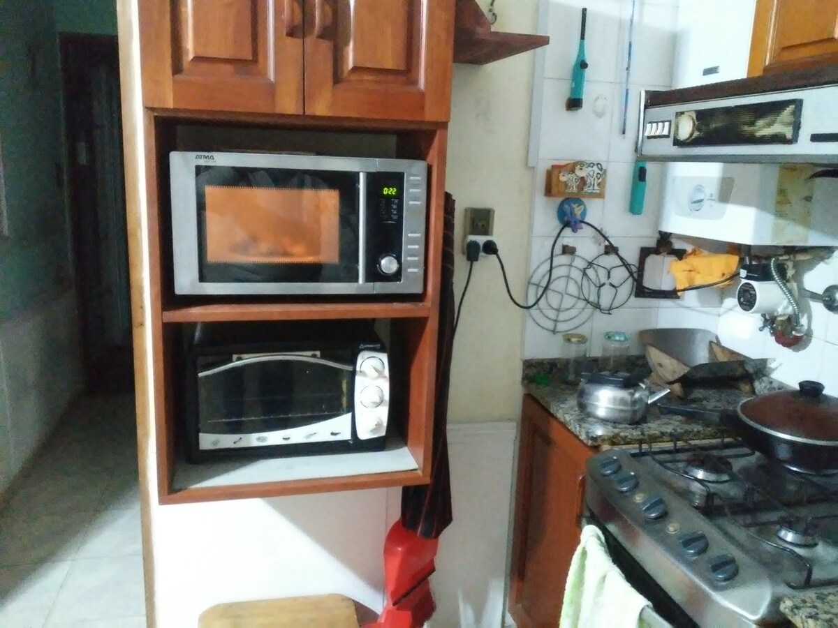 A kitchen area features a built-in microwave and a toaster oven positioned on a wooden shelf. Below, a traditional stove awaits use, surrounded by various kitchen utensils and equipment. The countertop displays pots and a small sink, contributing to a functional cooking space.