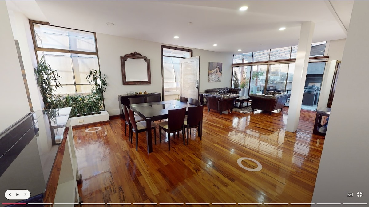 An open-plan living area is showcased, featuring polished wooden floors and large windows that allow natural light to illuminate the space. A dark wooden dining table with matching chairs is positioned beside a cozy seating arrangement. Indoor greenery adds a touch of nature to the elegant design.
