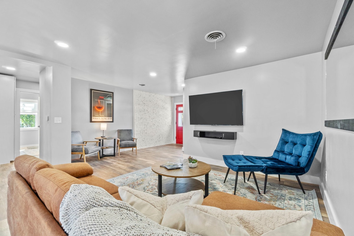 A bright living area features a spacious sofa and two accent chairs, arranged around a wooden coffee table. A large wall-mounted TV is positioned across from the seating. Natural light enters through nearby windows, enhancing the open feel of the room.