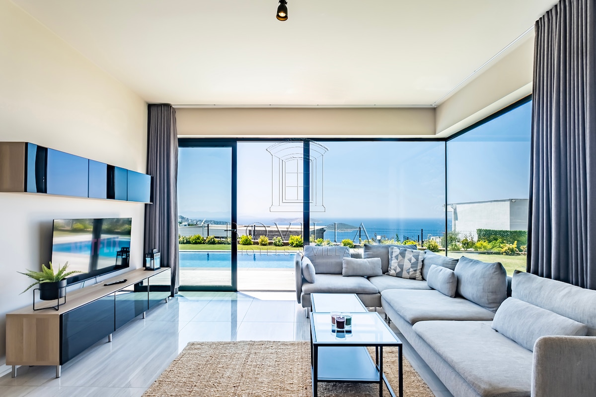 A bright living area is presented with large glass windows, offering unobstructed sea views. A comfortable sectional sofa arranged around a coffee table complements the sleek entertainment unit. Natural light fills the space, while soft curtains frame the windows, enhancing the open, airy feel.