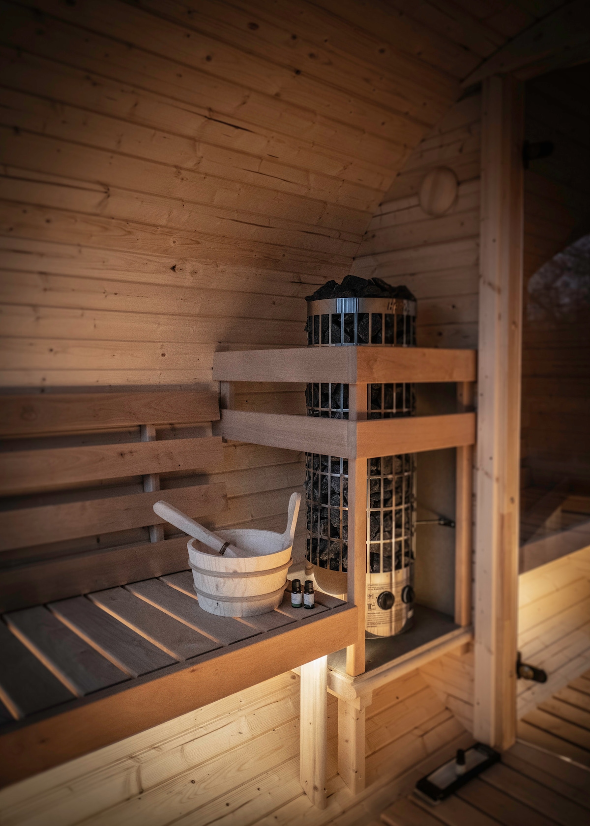 The interior of a sauna is shown, featuring warm wooden walls and benches. A wooden bucket and essential oil bottles are positioned nearby. A cylindrical heater is visibly integrated into the design, adding functionality to the relaxing space.