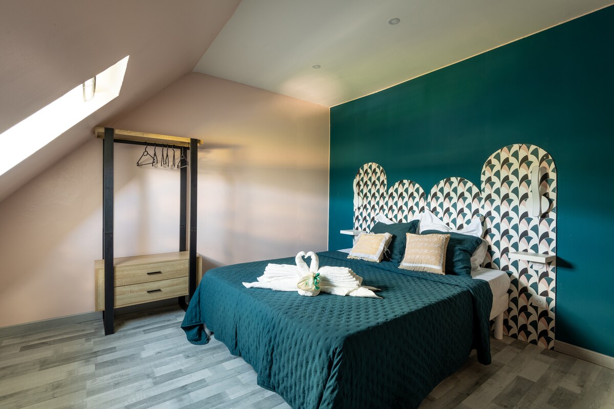A spacious bedroom features a vibrant teal accent wall with decorative elements above the inviting bed. Soft bedding is complemented by two decorative pillows. A minimalist wooden wardrobe stands on one side, providing storage and hanging space for clothing.