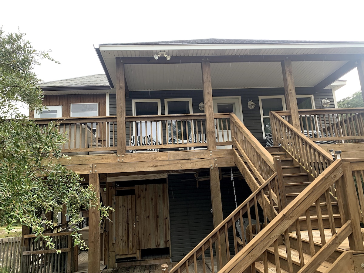 The exterior of the house features a spacious wooden deck with a series of steps leading up to the entrance. The porch is framed by railings and includes multiple seating options. Large windows allow natural light to brighten the interior, offering a welcoming view of the surrounding landscape.