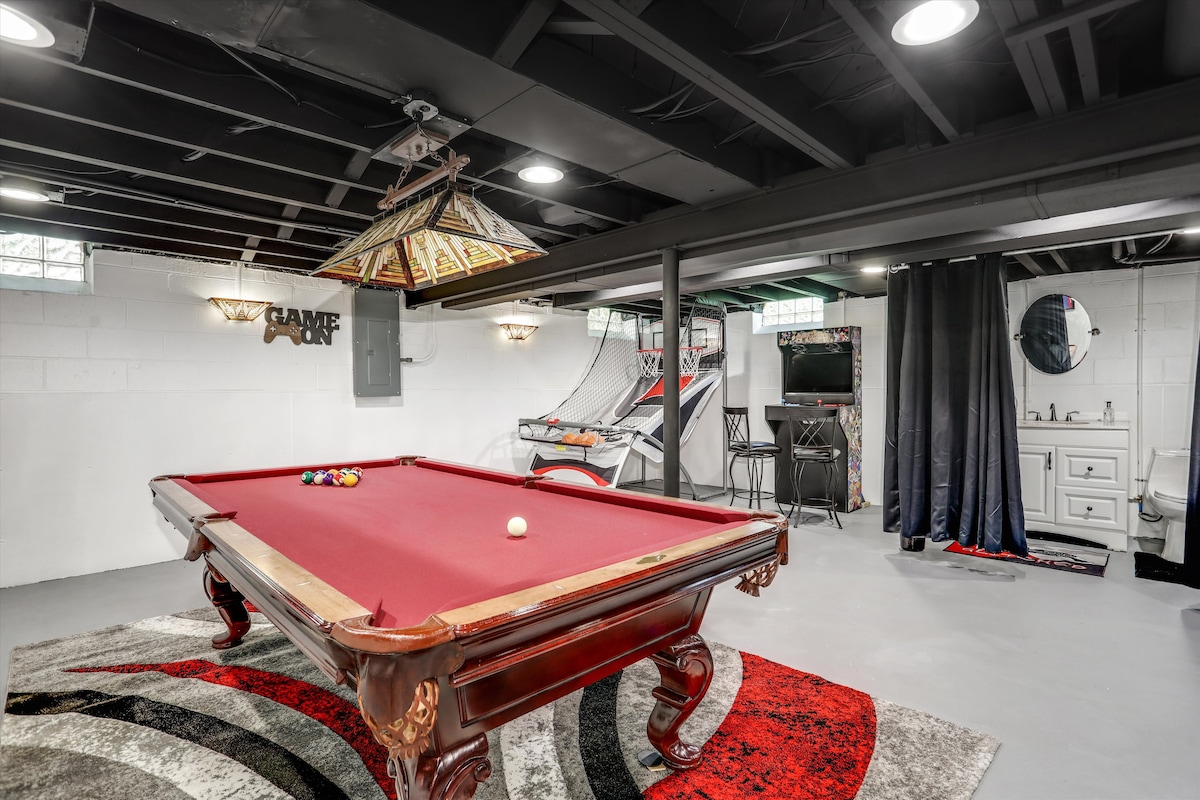 A game room features a vintage-style billiard table with a red felt surface positioned centrally. The space is equipped with an arcade basketball game in the background and a television mounted on the wall. Ambient lighting is provided by a decorative ceiling fixture.