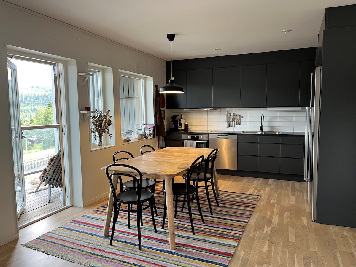 A modern kitchen is presented, featuring dark cabinetry and a light countertop. A wooden dining table with black chairs is centered on a colorful striped rug. Large windows allow natural light to fill the space, while an entrance door leads to an outdoor area.