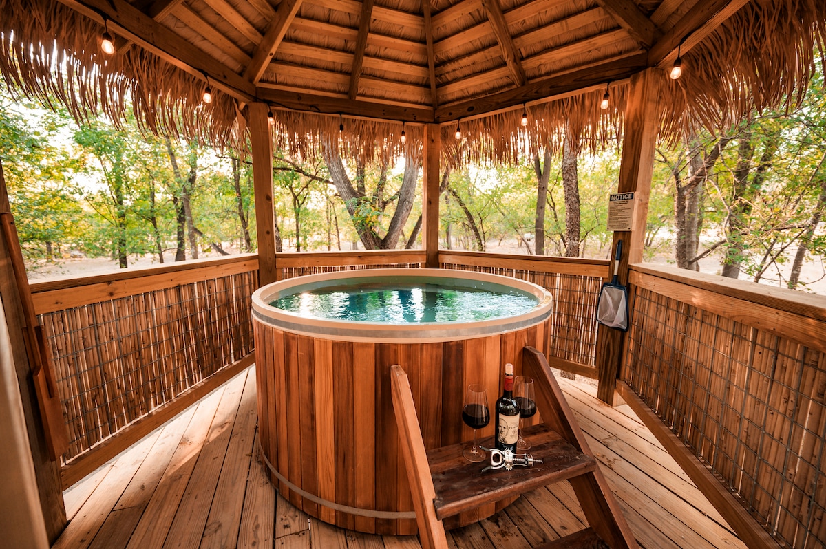 A wooden gazebo surrounds a large, circular hot tub, showcasing warm wooden tones and natural textures. Soft lighting is provided by string lights above. The area is surrounded by lush greenery, creating a peaceful retreat for relaxation.