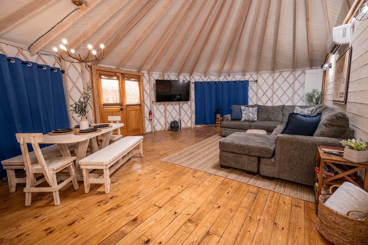 The open-concept living space is warmly inviting, featuring a comfortable sectional sofa and a large dining table with benches. Natural light fills the room through windows, enhancing the wooden floors and cozy atmosphere. A wall-mounted TV is positioned for group viewing.