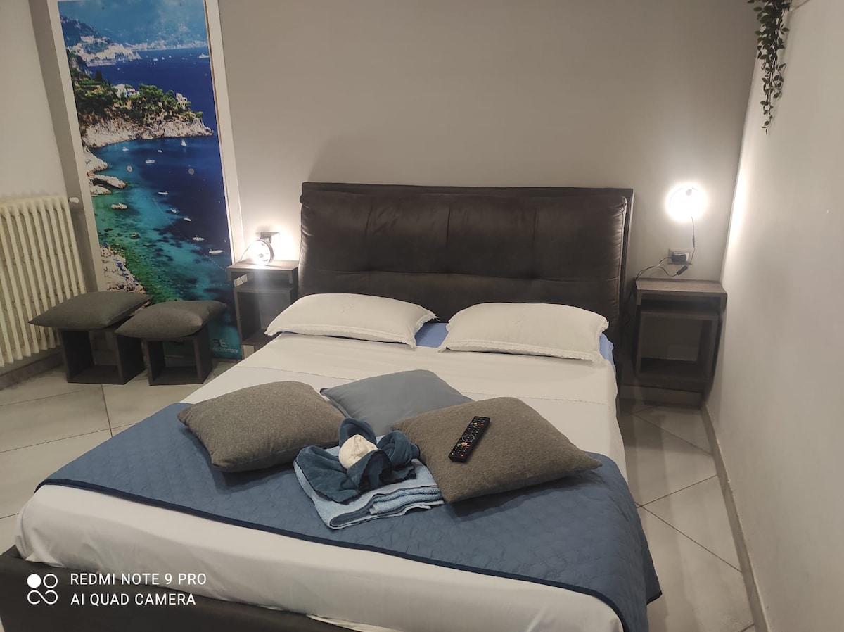 A spacious bed is positioned against a neutral wall, accompanied by two bedside tables featuring minimalist lamps. Decorative cushions lie on the bed, and a TV remote is casually placed atop a blanket. A coastal mural adds interest to the area.
