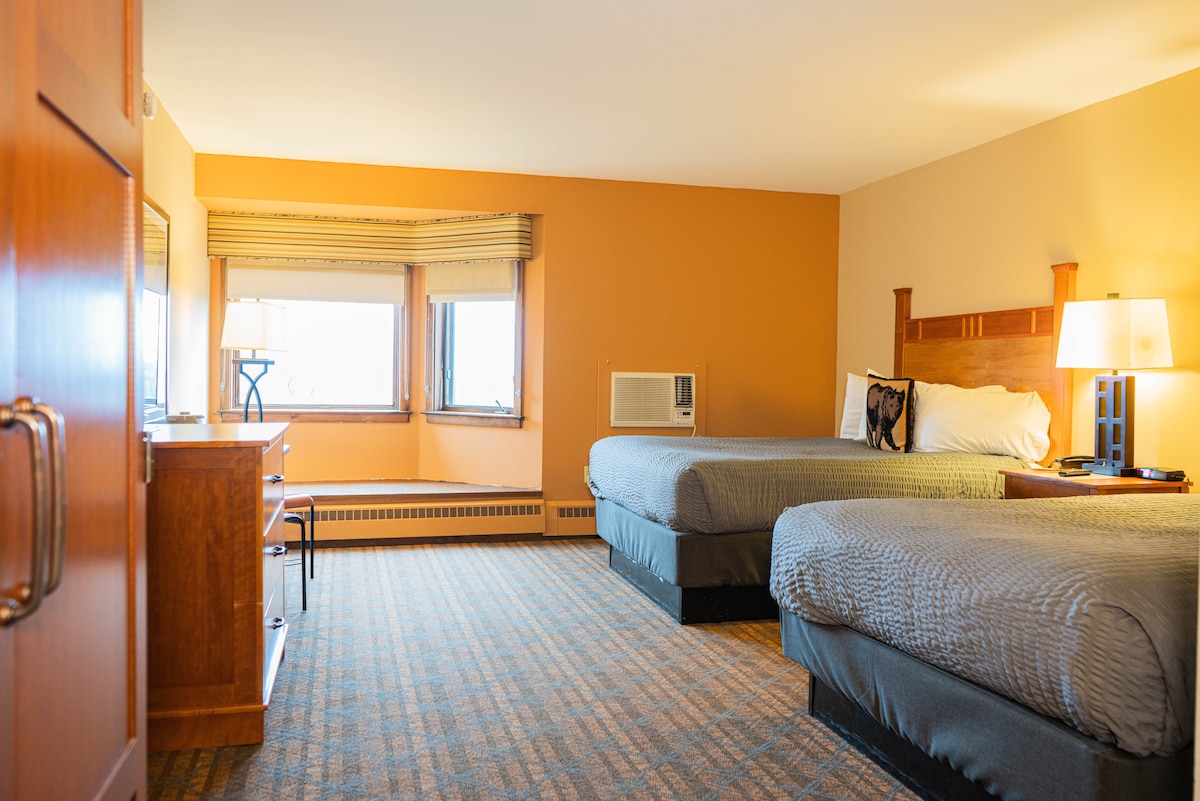 A well-lit guest room is equipped with two spacious beds and a wooden headboard. Natural light enters through multiple windows, illuminating the warm orange walls. A wooden dresser and lamp are positioned opposite the beds, while an air conditioning unit sits below one window.