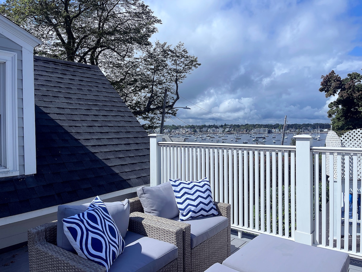 A cozy rooftop deck is equipped with a seating area featuring light-colored cushions and decorative blue and white pillows. The view includes a harbor surrounded by boats, with a partly cloudy sky and trees framing the scene.