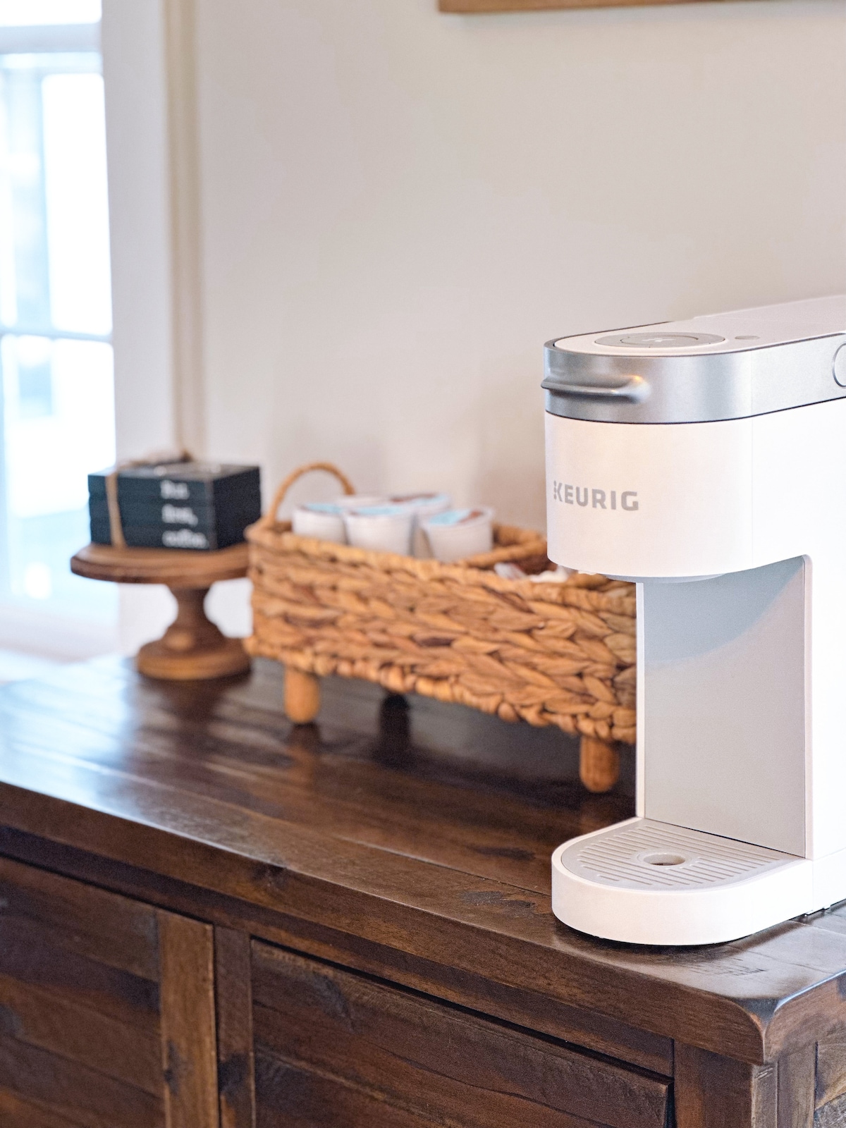 A Keurig coffee maker is positioned on a wooden console table. A woven basket nearby holds coffee cups, providing an organized and inviting coffee corner. Natural light enters through a nearby window, illuminating the space.