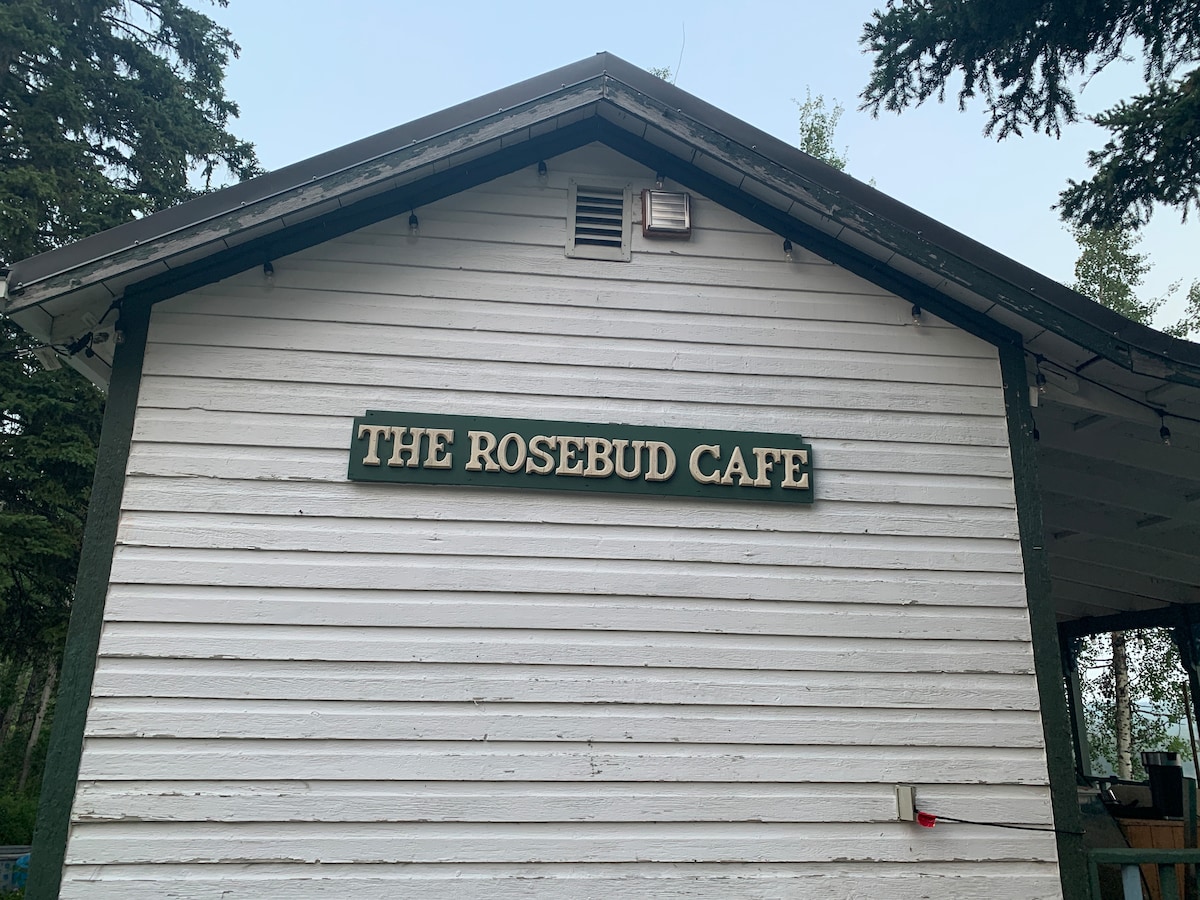 The exterior of the Rosebud Cafe is shown, featuring a wooden structure painted in white. A green sign with white lettering displays 'THE ROSEBUD CAFE.' Surrounding trees add to the natural setting.