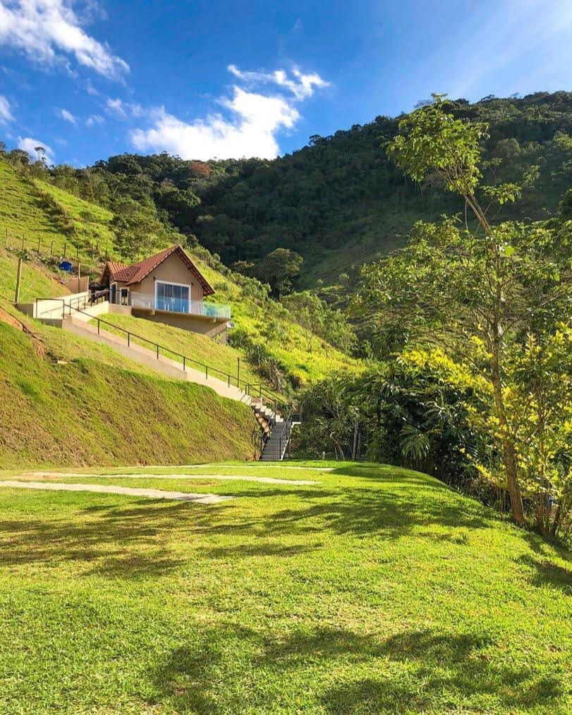 A charming chalet is positioned on a gently sloping hillside, surrounded by lush greenery. A pathway leads to the entrance, while a grassy area offers space for relaxation. The mountains rise in the background, and the sky is bright with scattered clouds.