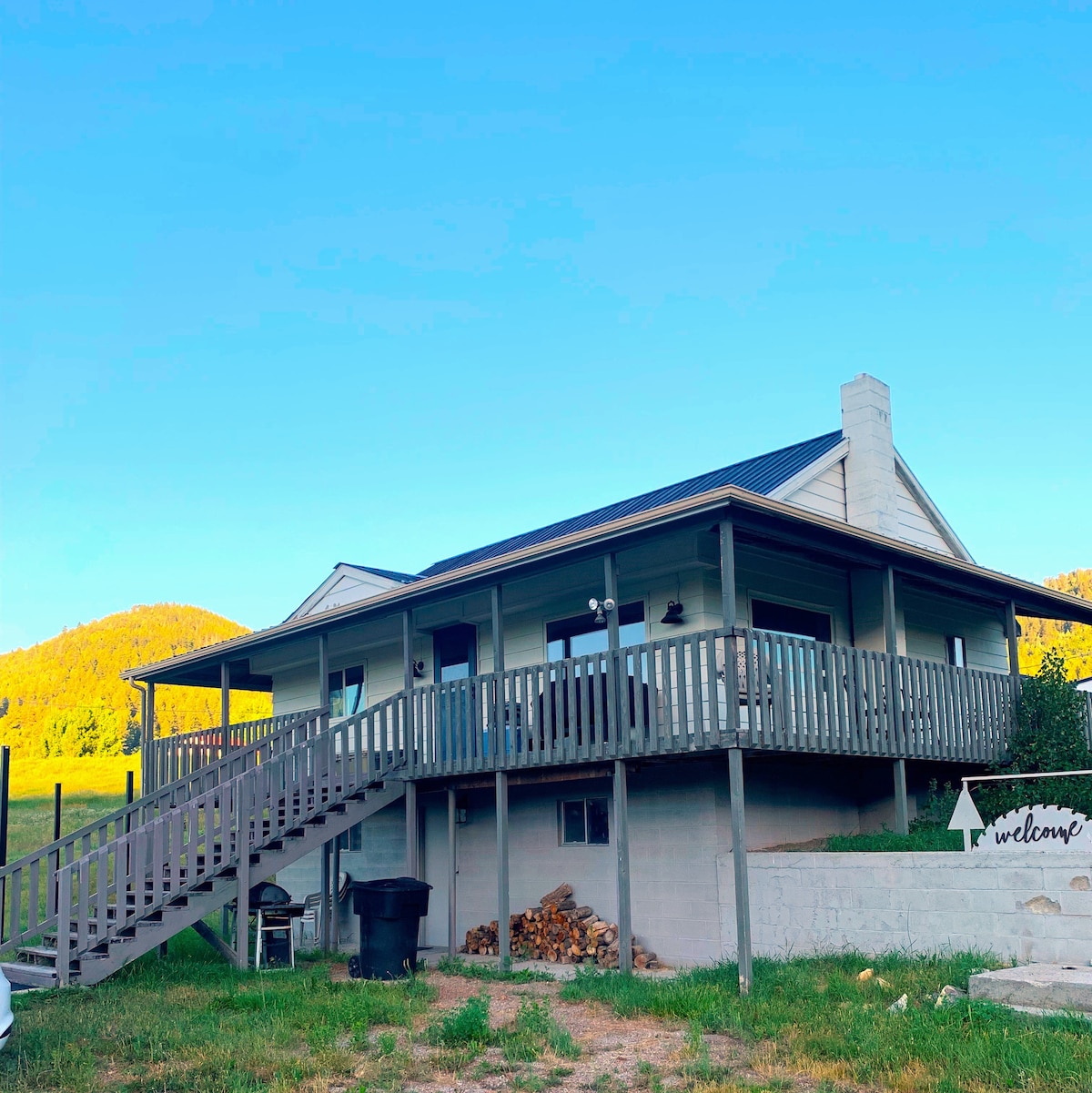 The farmhouse features a welcoming front porch with a staircase leading up to the entrance. A wooden deck wraps around the house, framed by grassy surroundings and gentle hills in the background. A sign indicating 'welcome' is positioned near the entrance.