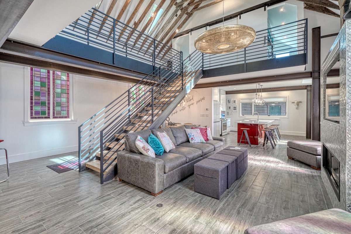 The spacious living area features an open floor plan with high ceilings, exposed wooden beams, and large windows. A comfortable sectional sofa, layered with colorful pillows, is positioned nearby a sleek dining space, while a staircase leads to the upper level.