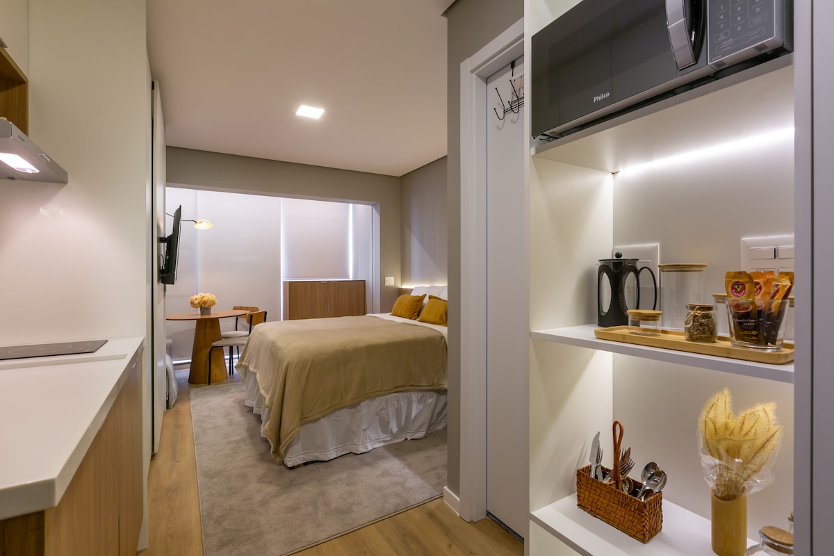 A modern studio apartment is shown, featuring a spacious bed with neutral-toned bedding. A kitchenette is visible on one side, equipped with essential appliances. Natural light filters in through large windows, brightening the room. A small dining table complements the open layout.