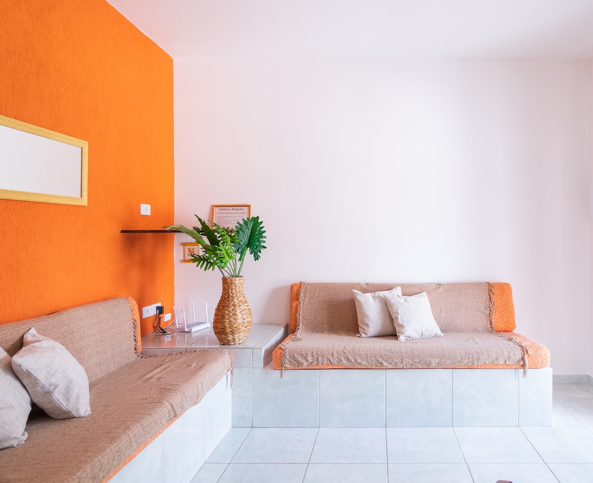 A bright living space features orange accent walls, enhancing the room's character. A woven basket holds greenery, while two benches with neutral fabric provide seating. A minimalist decor approach is evident, with light tones and a clean layout, contributing to an open and airy feel.