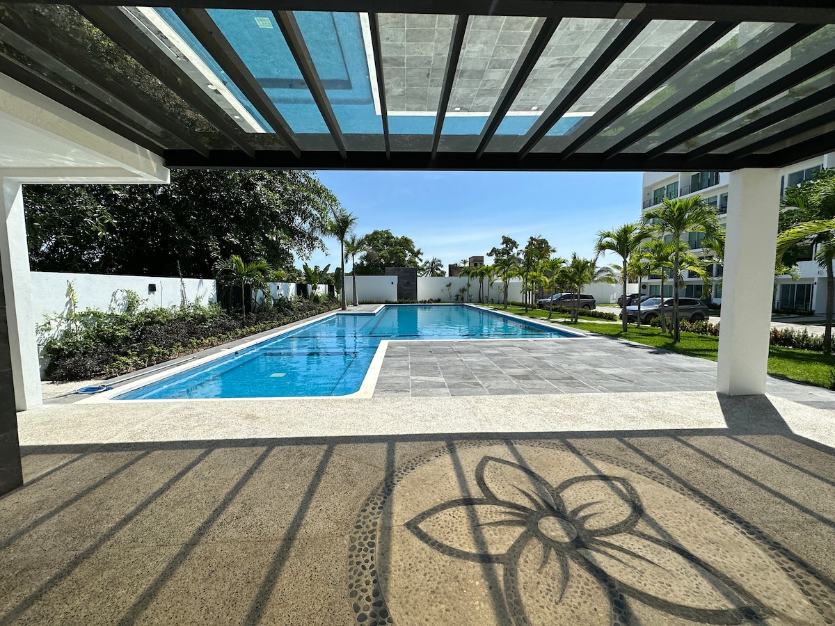 A large outdoor pool reflects the sky, surrounded by well-maintained gardens and palm trees. The area is shaded by a covered patio, providing a comfortable spot to relax and enjoy the view. The clean lines and open space invite leisurely afternoons.