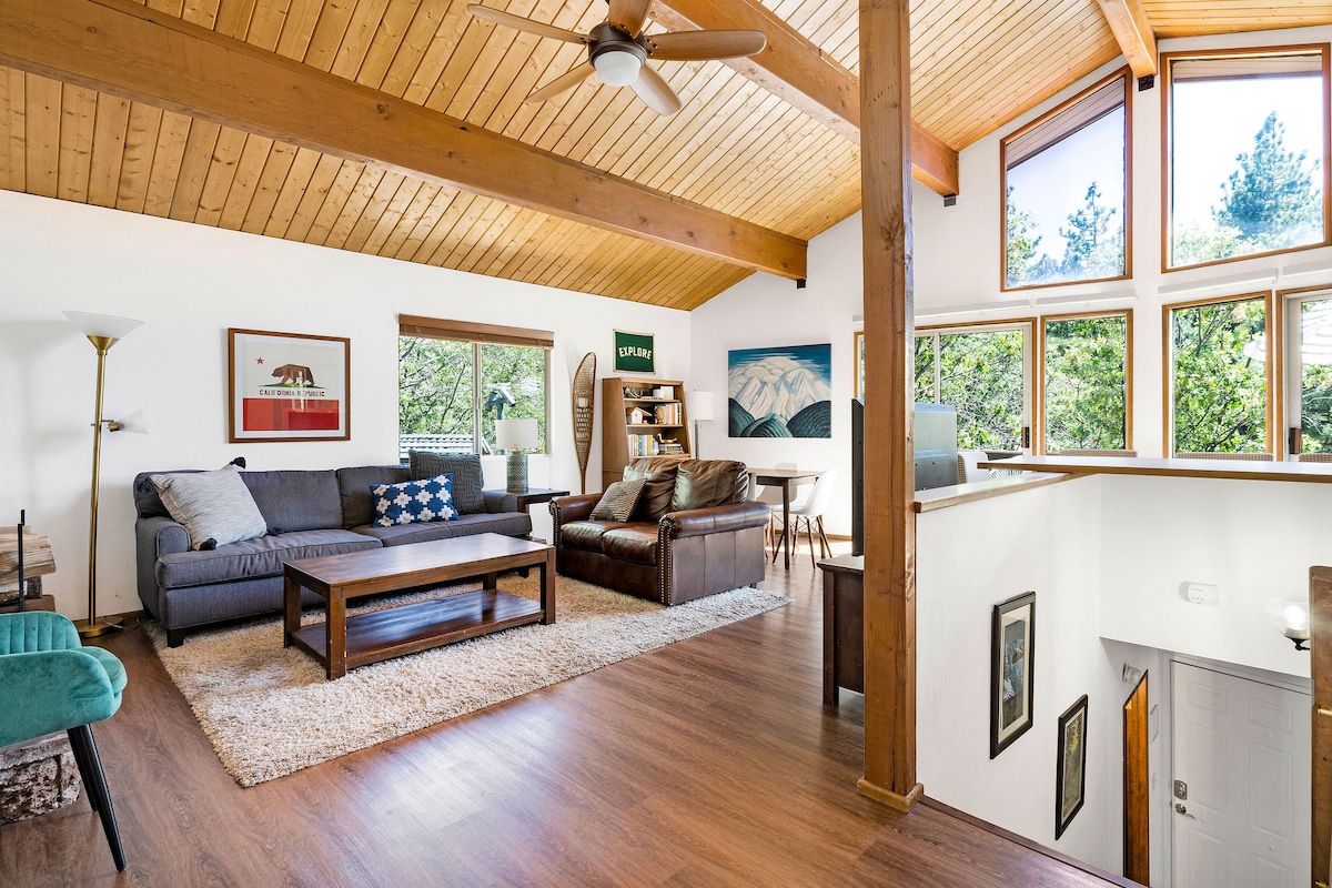 A spacious living area features a high vaulted ceiling with exposed wooden beams. A comfortable sofa and armchairs are arranged around a wooden coffee table, while large windows invite natural light and offer views of the surrounding trees.