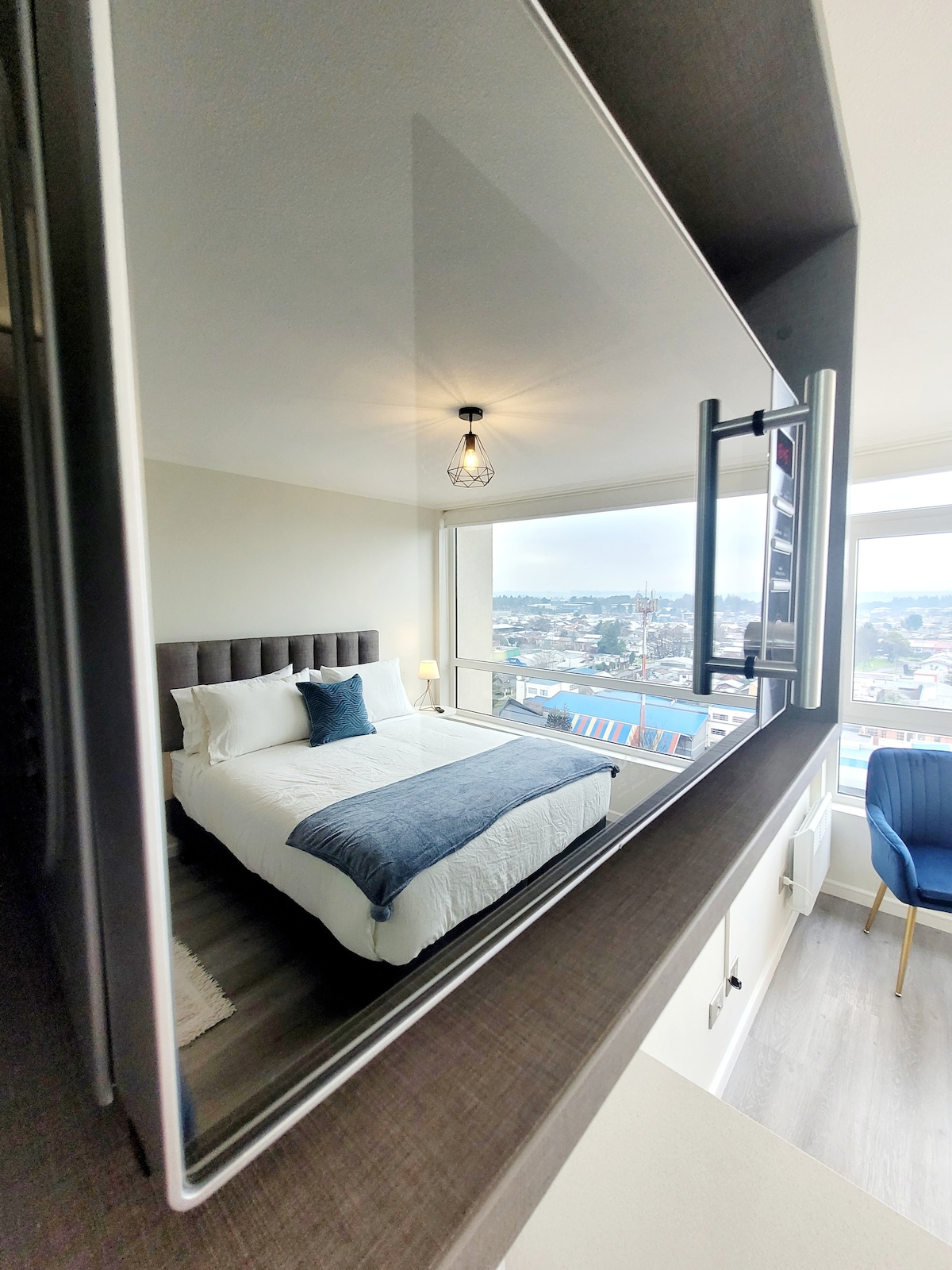 A view of a cozy bedroom is reflected in a modern mirror. The spacious bed is presented with a blue throw blanket and matching pillows. A nightstand with a lamp sits beside the bed, while a cozy chair is positioned near the window, offering distant city views.
