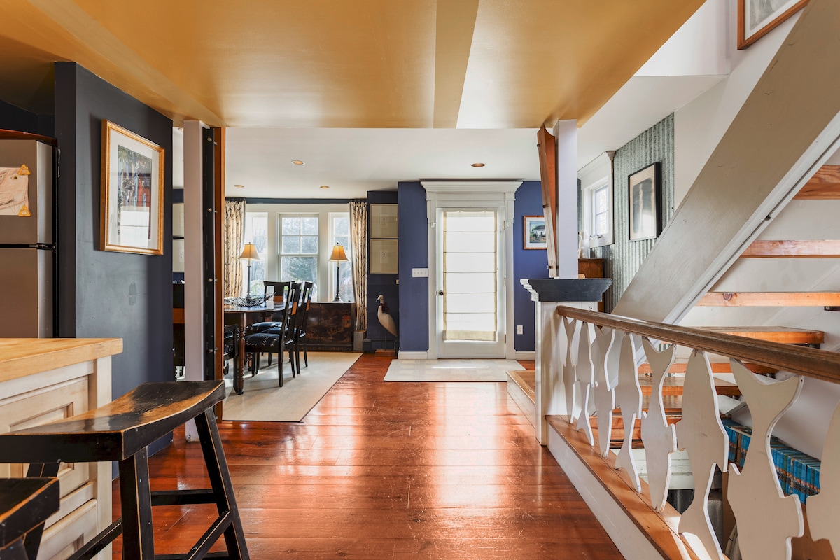 An open living space features rich wooden flooring and a combination of vibrant blue and neutral walls. A dining area is set against a large window, with a staircase visible in the background. Light is softly reflected off the space's high ceilings.