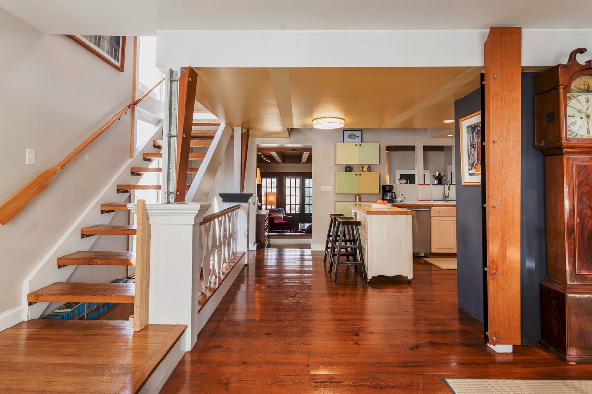 An open interior space is illuminated by natural light. A wooden staircase with a white railing leads to an upper level. The kitchen area features light-colored cabinetry, and bar stools are placed at the counter. Rich wooden flooring extends throughout the area.