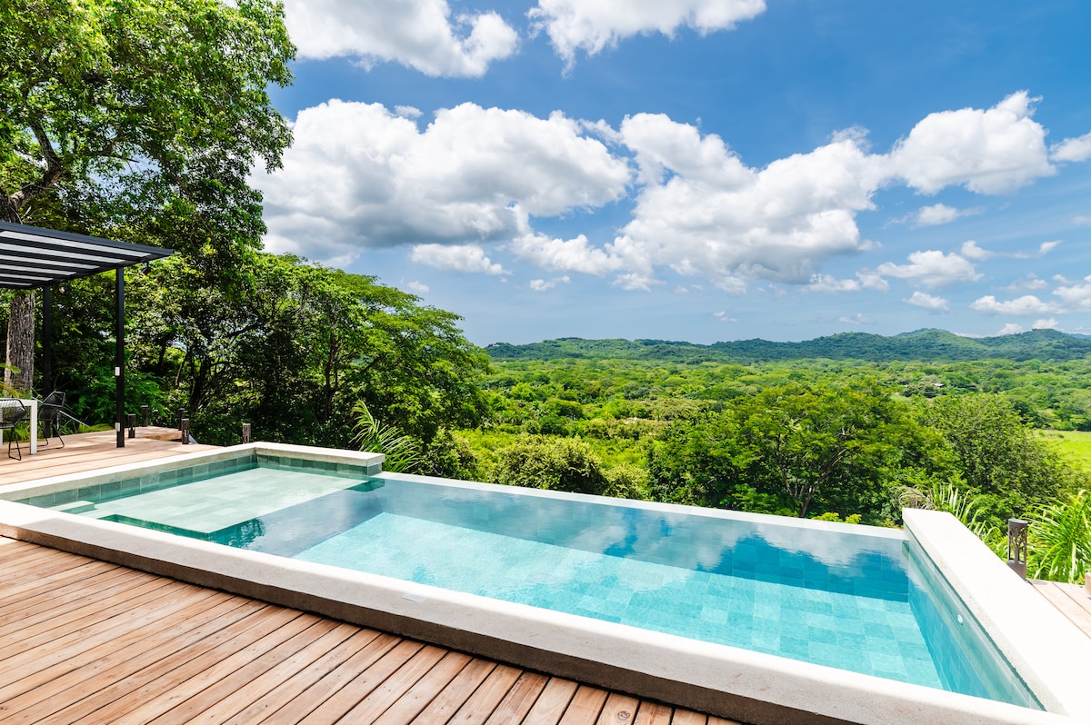 An infinity pool with a sleek design is surrounded by a wooden terrace. Lush greenery and rolling hills are visible in the background, under a bright blue sky dotted with clouds, creating a serene outdoor setting.