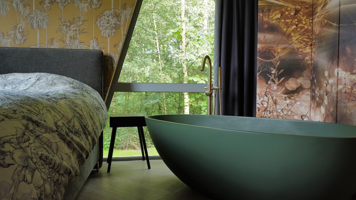 A serene bedroom features a spacious bed adorned with floral bedding. A modern freestanding tub in a soft green hue is positioned near a large window that showcases lush greenery outside. A minimalist black side table complements the calming atmosphere.