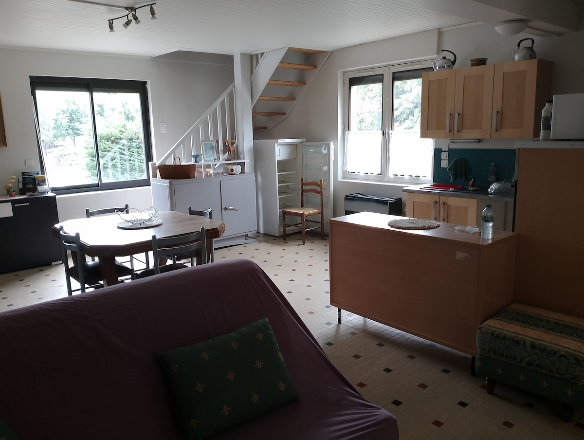 A spacious living area is showcased with a dining table surrounded by chairs and a kitchenette featuring essential appliances, including a refrigerator and microwave. A staircase leads to the upper floor, while large windows allow natural light to illuminate the space.