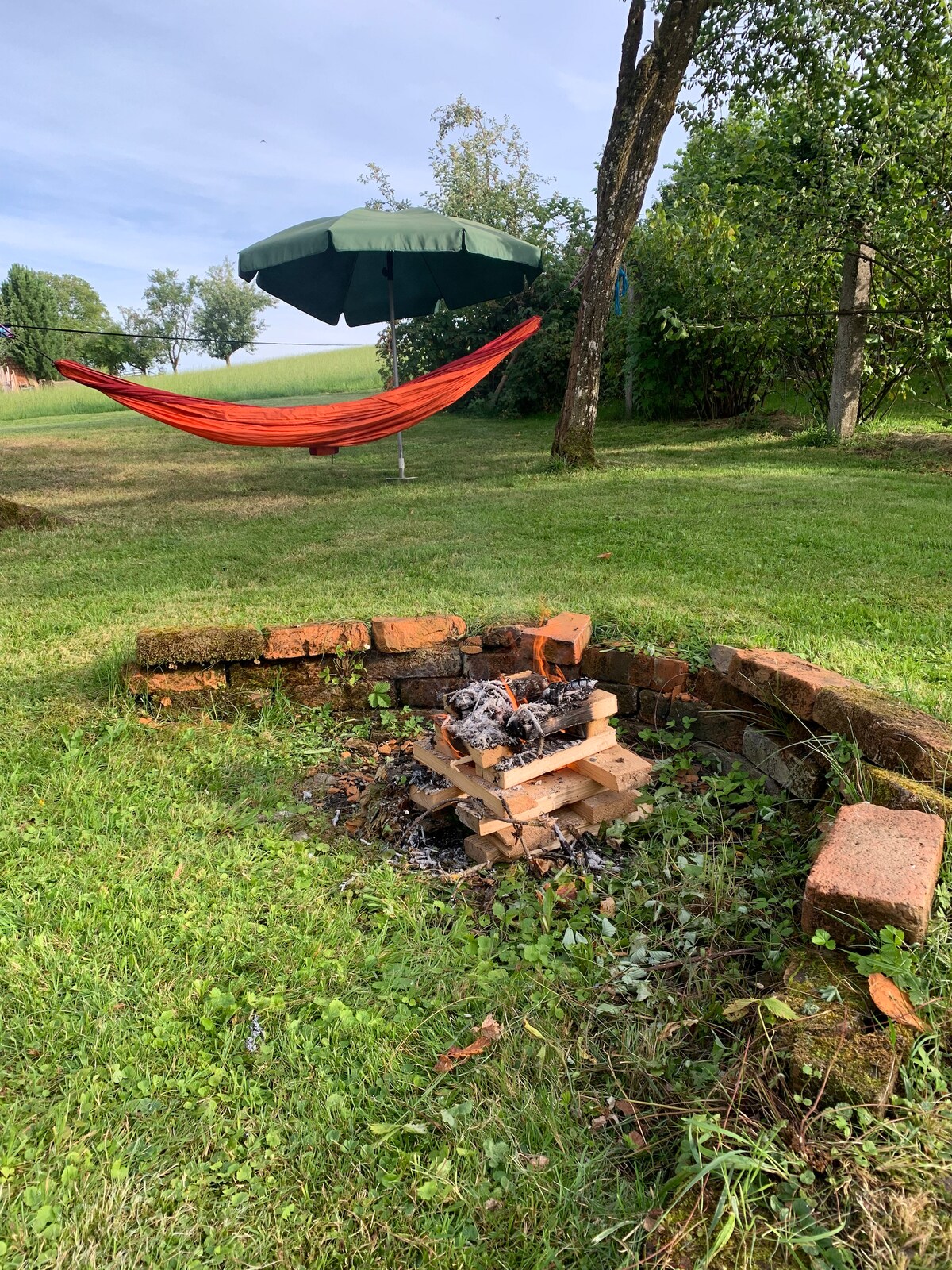 A fire pit, framed by a circular stone border, shows remnants of charred wood and ashes. Nearby, an orange hammock is suspended between two trees, complemented by a large green umbrella providing shade. The grassy area features scattered trees in the background.