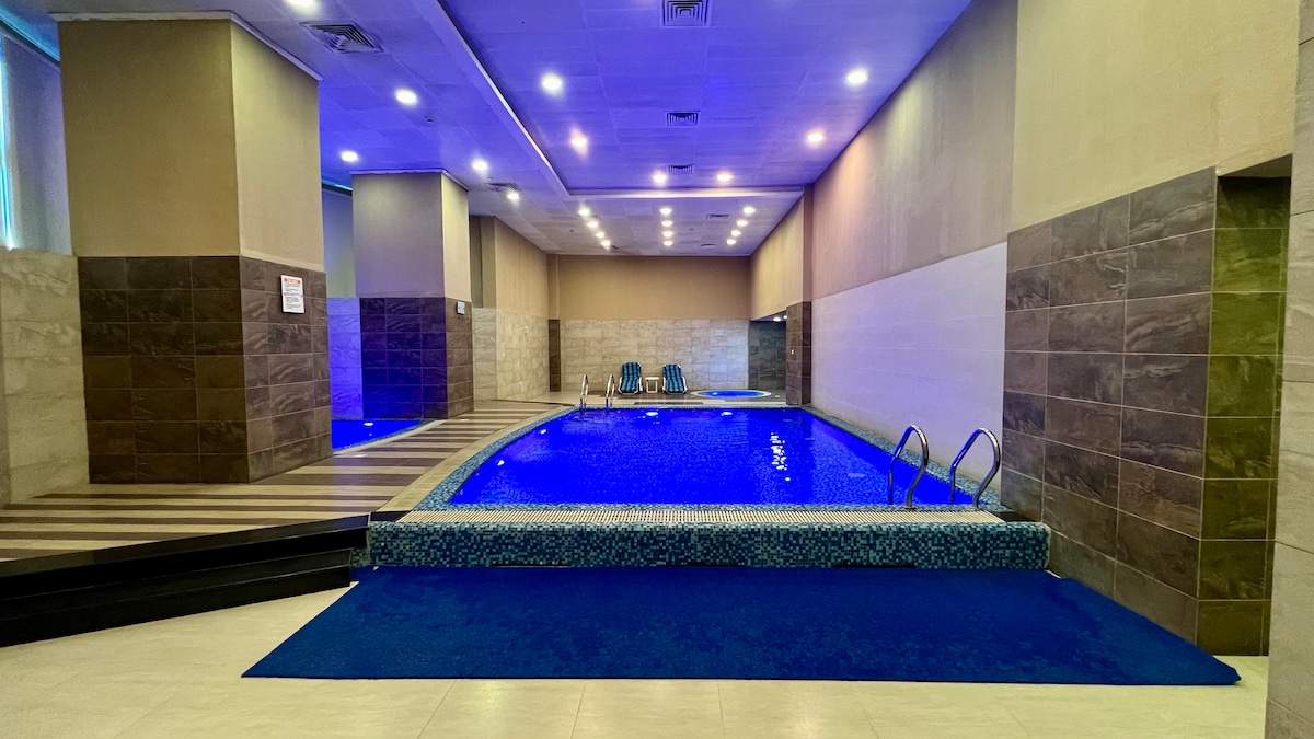 The indoor swimming pool is illuminated with calming blue lights, surrounded by tiled walls. A spacious area features lounge chairs positioned nearby, creating a serene environment for relaxation. Steps lead into the water, providing easy access for guests.