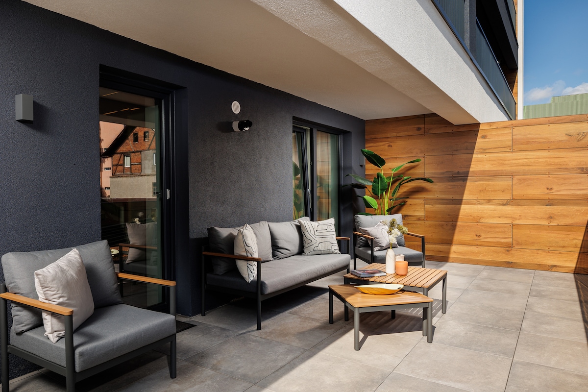 A spacious terrace features modern outdoor furniture, including a comfortable sofa set and a wooden coffee table. A vibrant green plant adds a touch of nature, while wooden panels provide a sense of privacy. The area is bathed in natural light from the nearby windows.