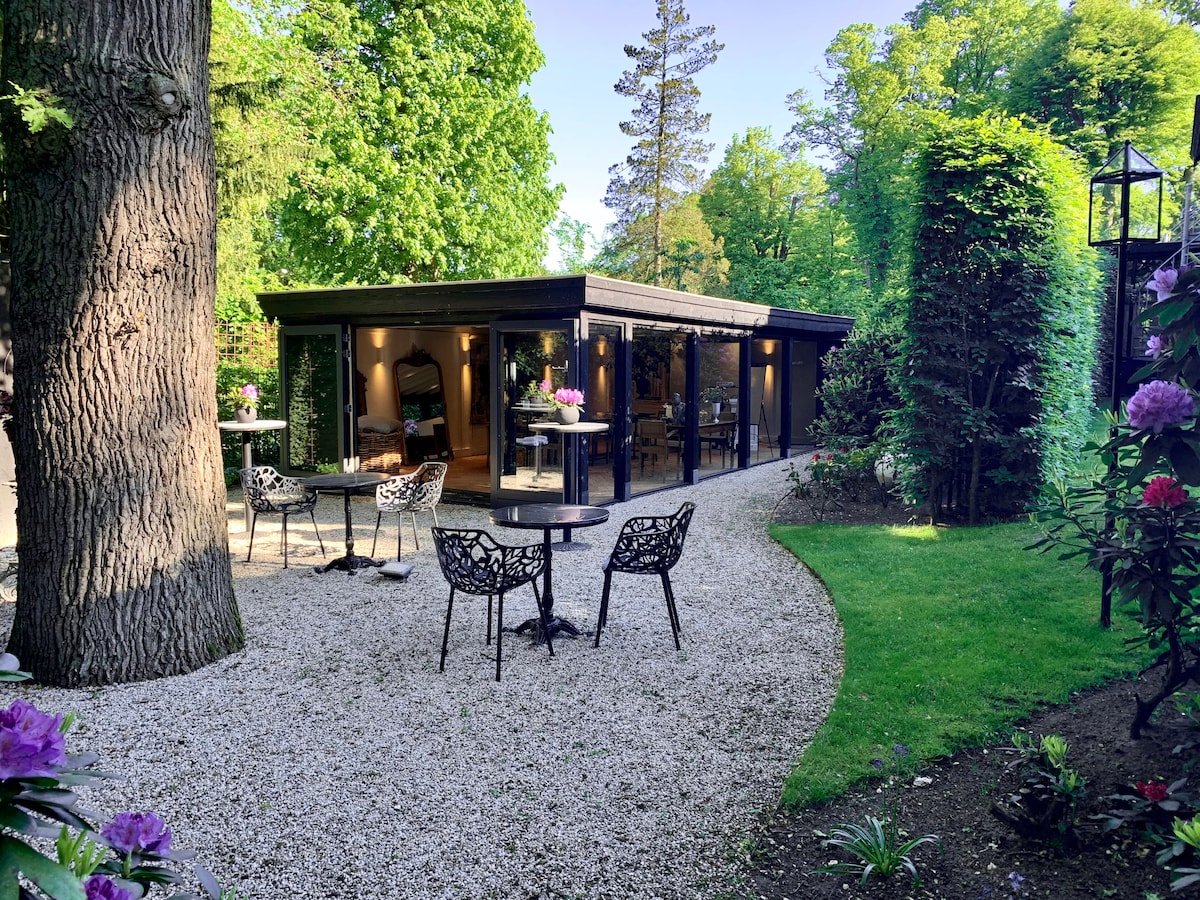 A modern studio cabin is set within a lush garden, surrounded by trees. Outdoor seating arrangements with small tables and chairs are present on pebbled pathways, providing a tranquil setting. Natural greenery frames the pathway leading to expansive glass doors of the studio.