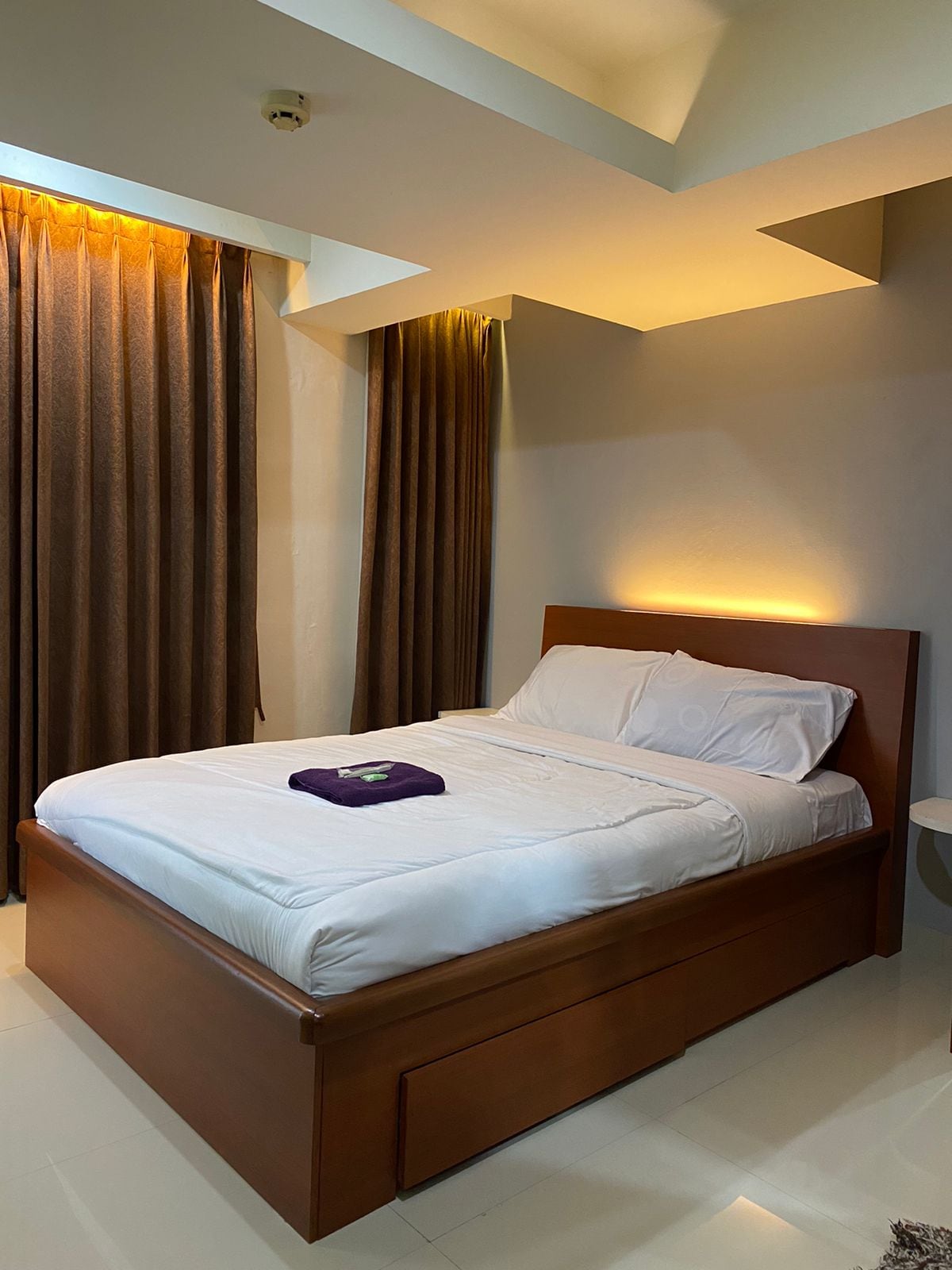 A well-lit bedroom features a spacious bed with crisp white linens and a folded towel placed on the blanket. Dark curtains frame the windows, and soft lighting is present above the headboard, creating a serene atmosphere.