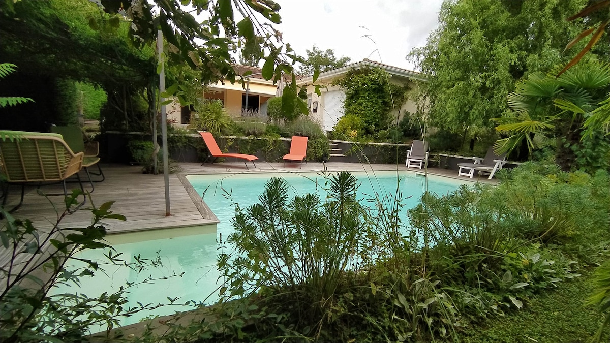 A serene swimming pool is surrounded by lush greenery and tropical plants. Two sun loungers in vibrant orange face the water, while additional seating is visible nearby. The house is partially seen in the background, nestled within the garden's natural beauty.