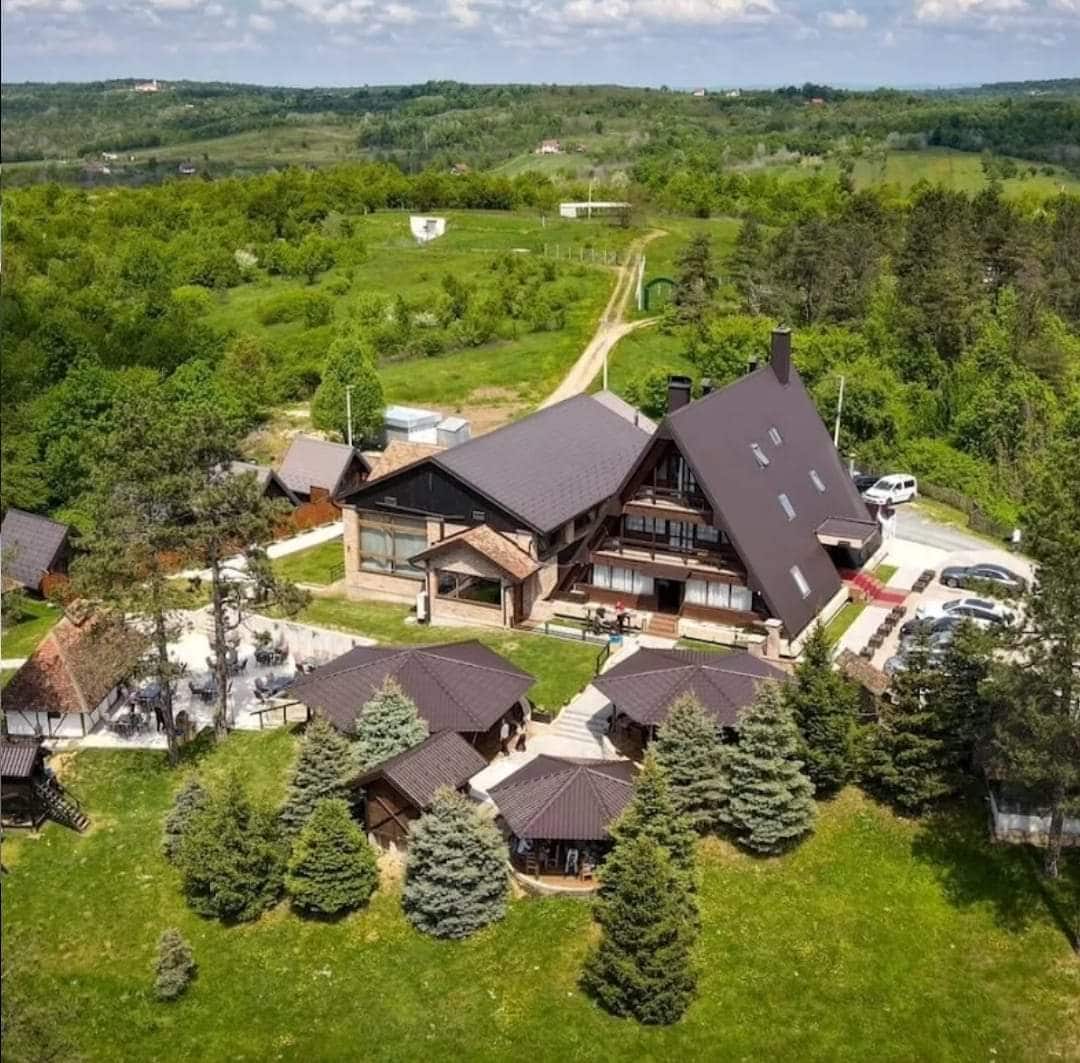 An aerial view presents a spacious property surrounded by lush greenery. The main building features a modern design with a sloped roof, complemented by several smaller structures with matching roofs. A pathway leads through the landscape, enhancing the tranquil outdoor setting.