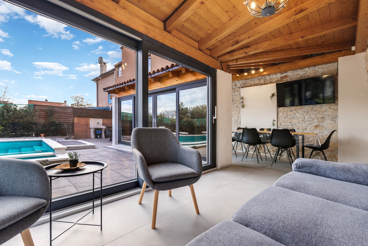 An inviting living area features large glass doors that open to a private pool and patio. Natural light fills the space highlighting the wooden ceiling and stone walls. Two comfortable armchairs are positioned beside a cozy sofa, with a dining area visible in the background.