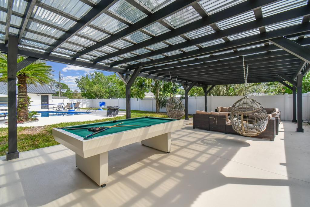 An outdoor covered lounge area features a billiard table and a comfortable seating arrangement. Two hanging chairs are suspended nearby. The space is surrounded by greenery and offers a view of the pool, creating a relaxing atmosphere.