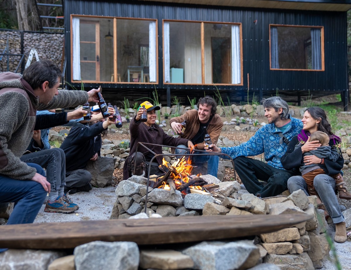 A gathered group enjoys a fire pit surrounded by stone seating. Warm light from the flames creates a cozy atmosphere. The backdrop features a modern cabin with large windows revealing a natural setting. Natural materials and greenery complement the space.