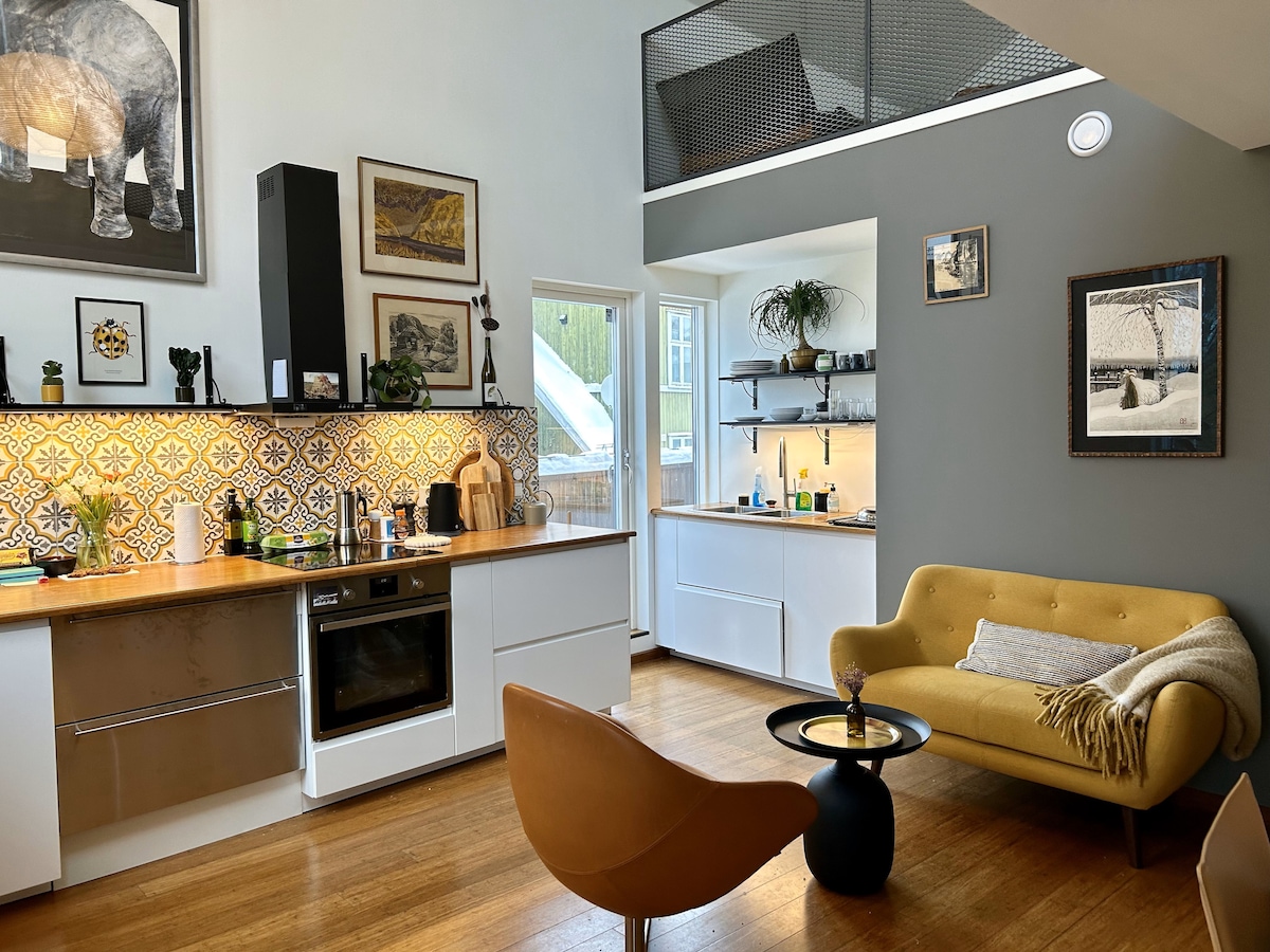 A modern kitchen and living area are depicted, featuring white cabinetry, a wooden countertop, and patterned tile backsplash. A yellow sofa and a small black table are positioned in the living space, complemented by a stylish chair. Artwork adorns the walls, enhancing the inviting atmosphere.
